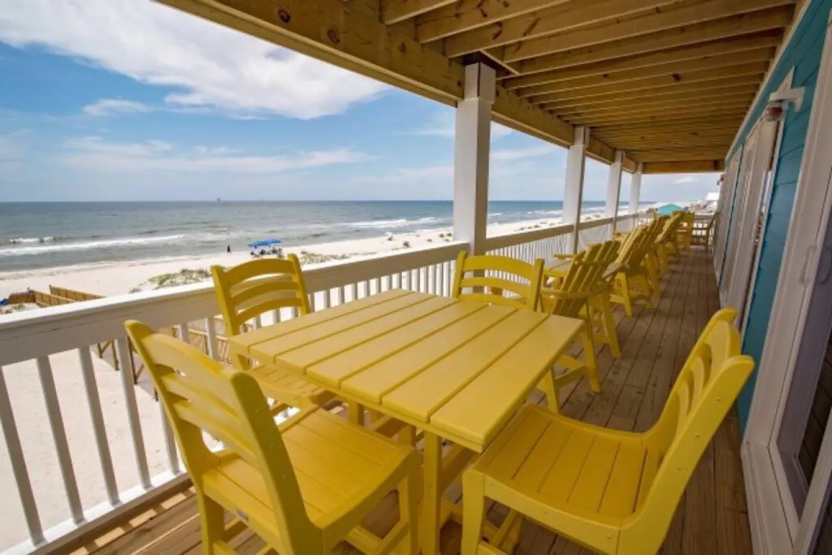 An exterior view showcasing a yellow dining table on a wooden deck overlooking the ocean and beach. The setting is ideal for outdoor dining, relaxing with friends, and enjoying stunning sea breezes and sunsets.