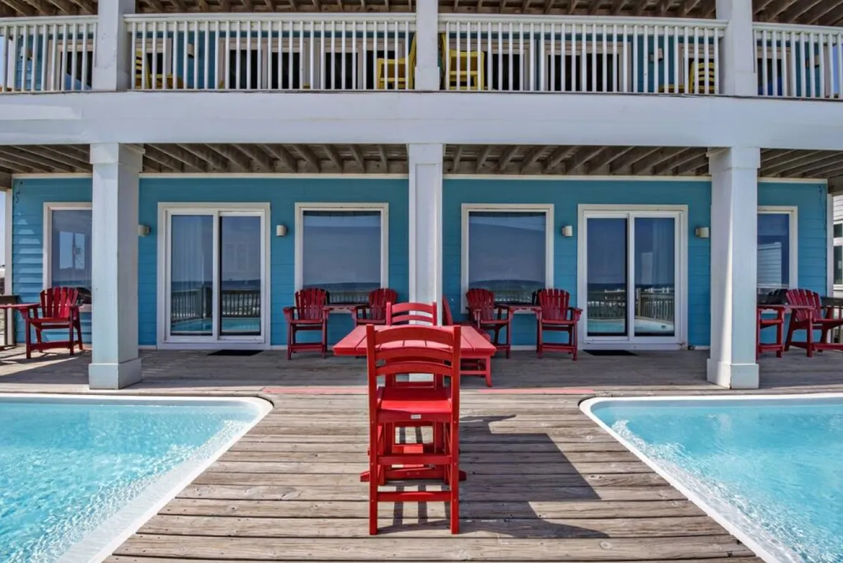 An inviting scene featuring a red outdoor dining set on a wooden deck, adjacent to a sparkling swimming pool surrounded by blue exterior walls and adirondack chairs. The area is sun-drenched and ready for swimming, dining, or lounging.