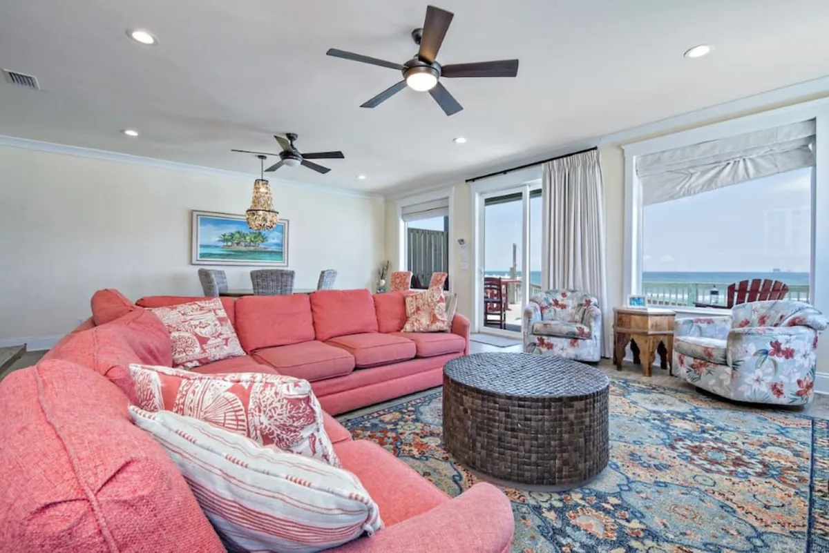 An inviting open living area with a large pink sectional sofa, woven coffee table, and floral accent chairs facing expansive ocean views through oversized windows. The relaxed, communal atmosphere is perfect for family and friends to unwind or celebrate together.