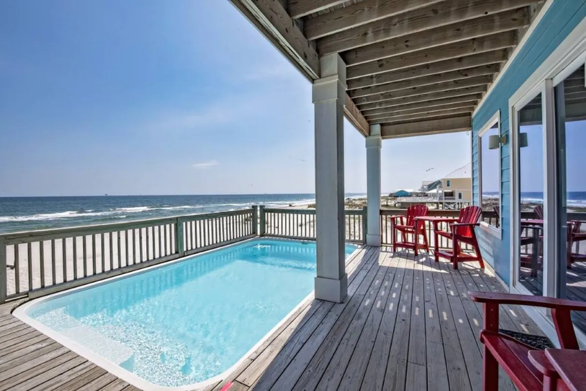 A sparkling beachfront pool sits on an elevated deck, overlooking the vast ocean. Red chairs and a spacious deck area invite guests to lounge, swim, and soak in the sea views. The setting transitions easily from playful daytime swims to tranquil sunset moments.