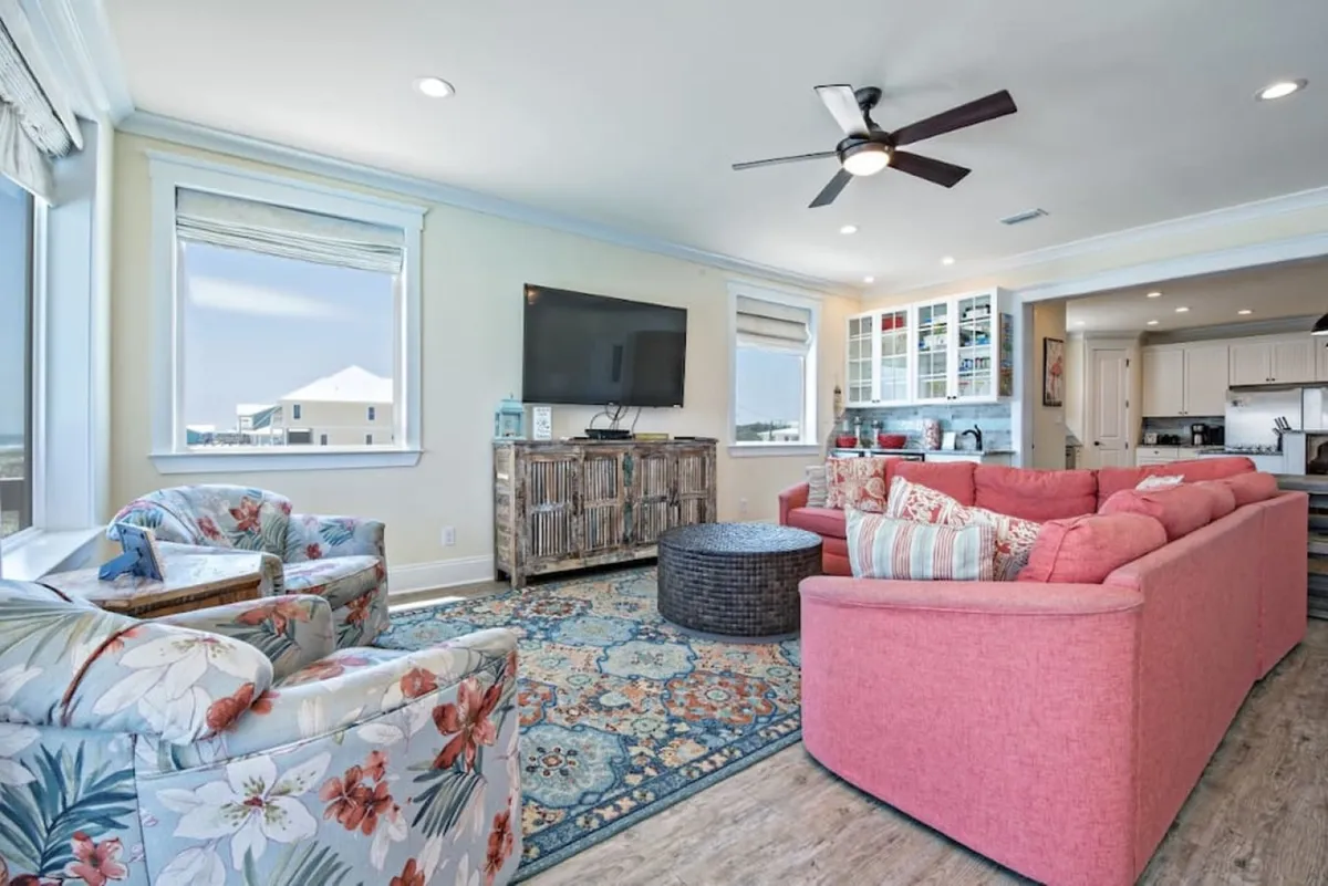 A casual living room with a pink sectional sofa, floral armchairs, a coffee table, and a television, all set within an inviting, homey atmosphere. The space is designed for relaxing, socializing, and intimate family gatherings, offering comfort and a sense of togetherness.