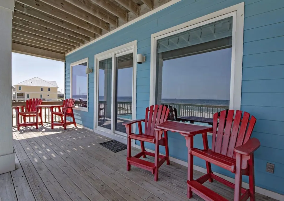 A welcoming outdoor deck featuring red Adirondack chairs faces the ocean, with large windows and blue exterior walls in the background. The space is set up for socializing, enjoying sunrises, or hosting an intimate gathering while taking in breathtaking views.