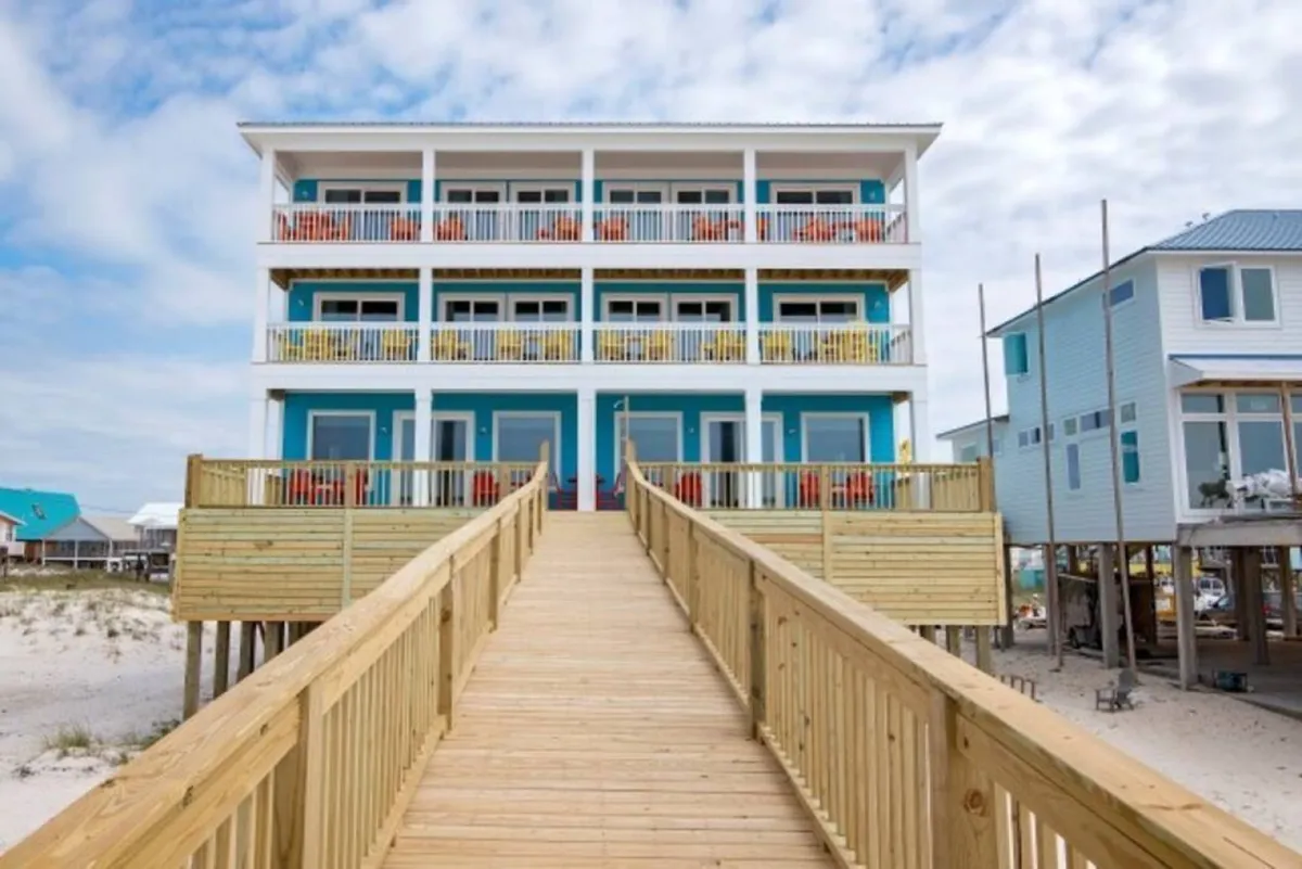 A cheerful exterior shot displaying a wooden walkway leading up to colorful balconies on a blue beach house. The inviting outdoor space is perfect for hosting or relaxing, with easy access to the outdoors and scenic views.