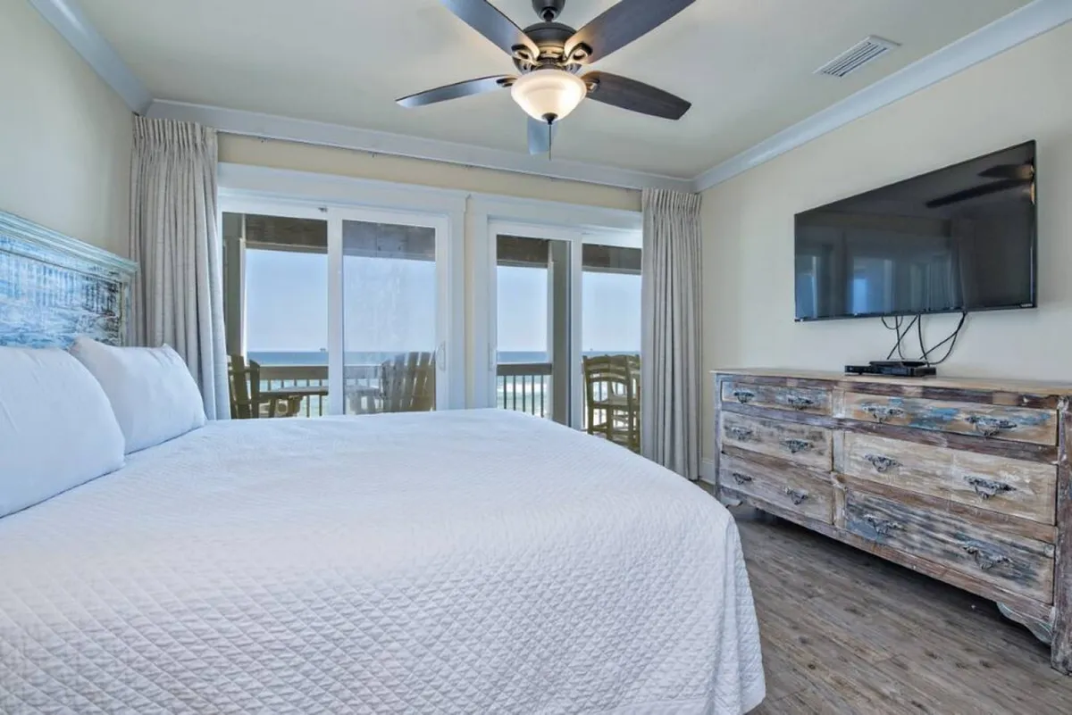 A serene king suite featuring a king-size bed with crisp plush linens, large windows that open to a balcony with ocean views, a wooden dresser, and a ceiling fan. The tranquil color palette and blackout shades create a peaceful, restful environment.