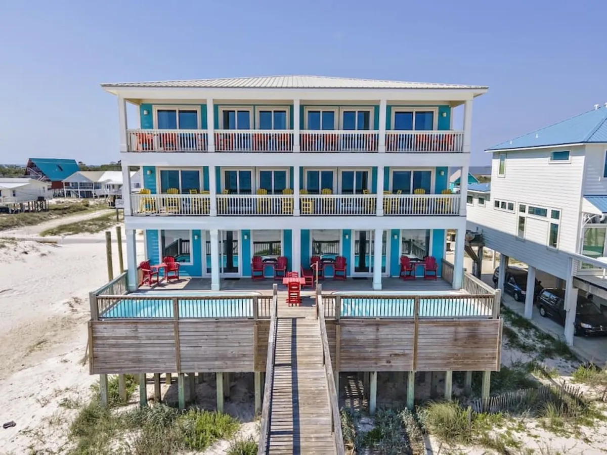 The image showcases a stunning three-story beach house duplex right on the sugar-white sands of Gulf Shores. Multiple balconies overlook private beachfront pools surrounded by red lounge chairs. A wooden walkway leads directly from the sand to the property, inviting guests to step from the beach into luxury. Spacious decks offer plenty of space for relaxation, with the serene Gulf of Mexico as a breathtaking backdrop.