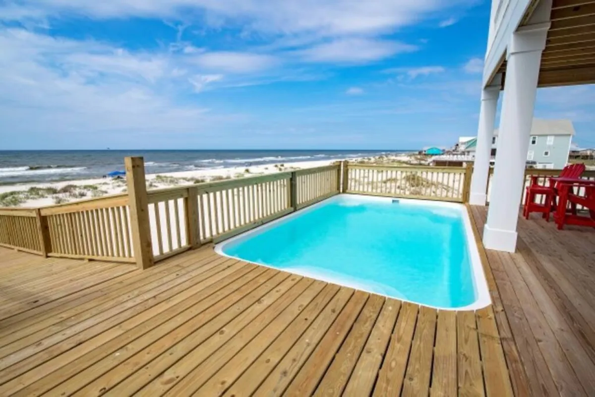 A private beachfront pool sits adjacent to a beach house, with a wooden deck furnished with red Adirondack chairs. Beyond the pool, uninterrupted ocean views create a luxurious and serene backdrop, emphasizing both relaxation and family fun in a private setting.