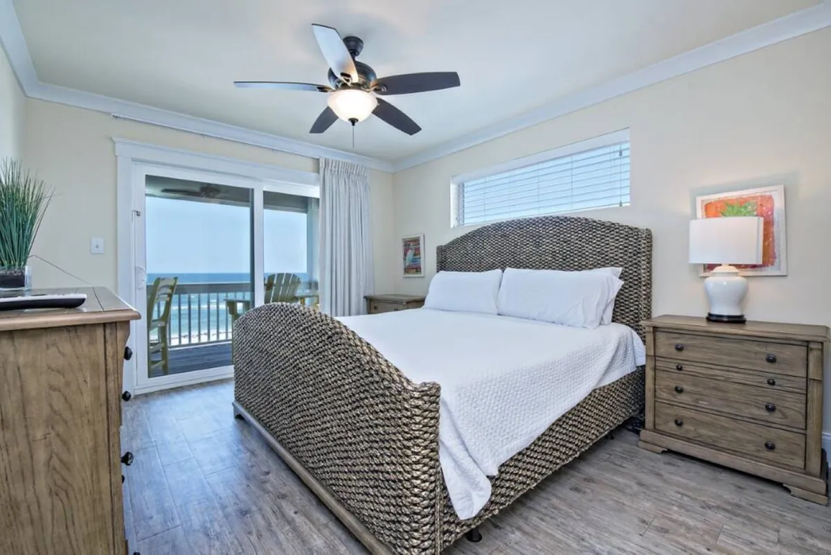 A serene bedroom with a woven textured bed frame faces a large glass sliding door that opens to a balcony with an ocean view. Natural light pours in, highlighting the wooden dresser with plant decoration and the cozy bedside tables with lamps.
