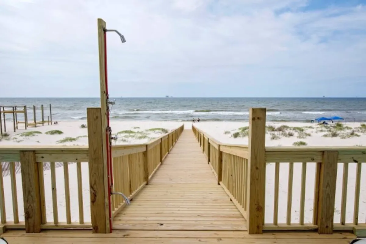 A wooden boardwalk/pathway stretches toward a quiet beach, with an outdoor shower visible for rinsing off after swimming. Blue beach umbrellas dot the distance, and the sparse dunes provide a natural, peaceful buffer from the outside world.