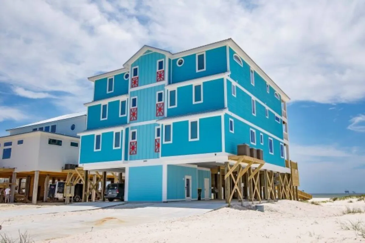 A vibrant blue three-story beach house is elevated on stilts above sandy dunes, with red and white decorative shutters and a cheerful, clear sky overhead. The surrounding area features open sandy spaces and a sense of privacy, encapsulating the spirit of coastal leisure and group gatherings.