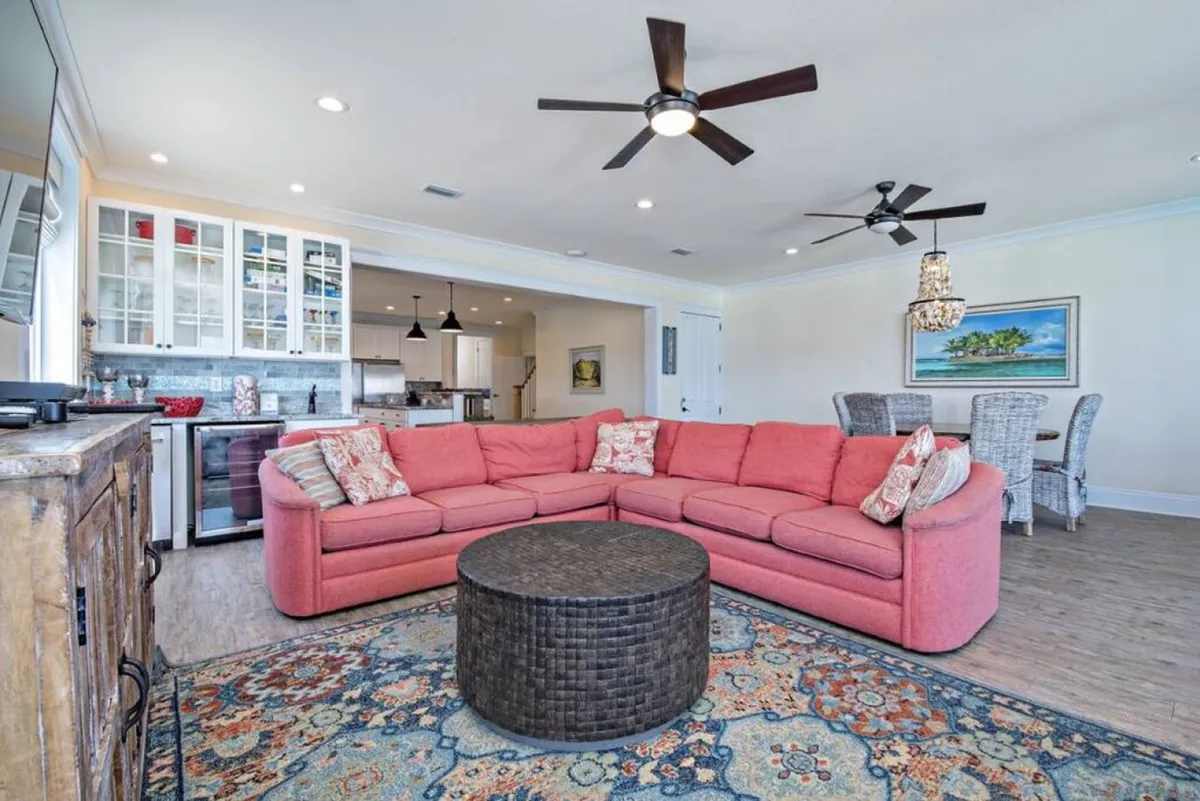 A bright, open-plan living and dining area features a large coral sectional sofa, a round woven coffee table, and an adjacent kitchen with white cabinetry and a dining area with wicker chairs and coastal artwork. The space is airy, inviting, and designed for relaxation and group gatherings.