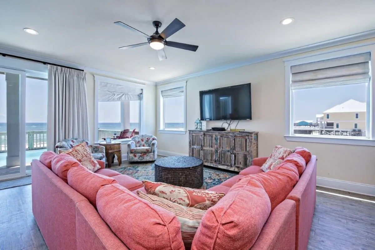 A spacious, bright living area featuring a large coral-pink sectional sofa, wall-mounted flat-screen TV, and a round woven coffee table. Floor-to-ceiling windows and a sliding glass door reveal panoramic Gulf views, with light streaming in and a seamless connection to the beachfront deck just beyond.