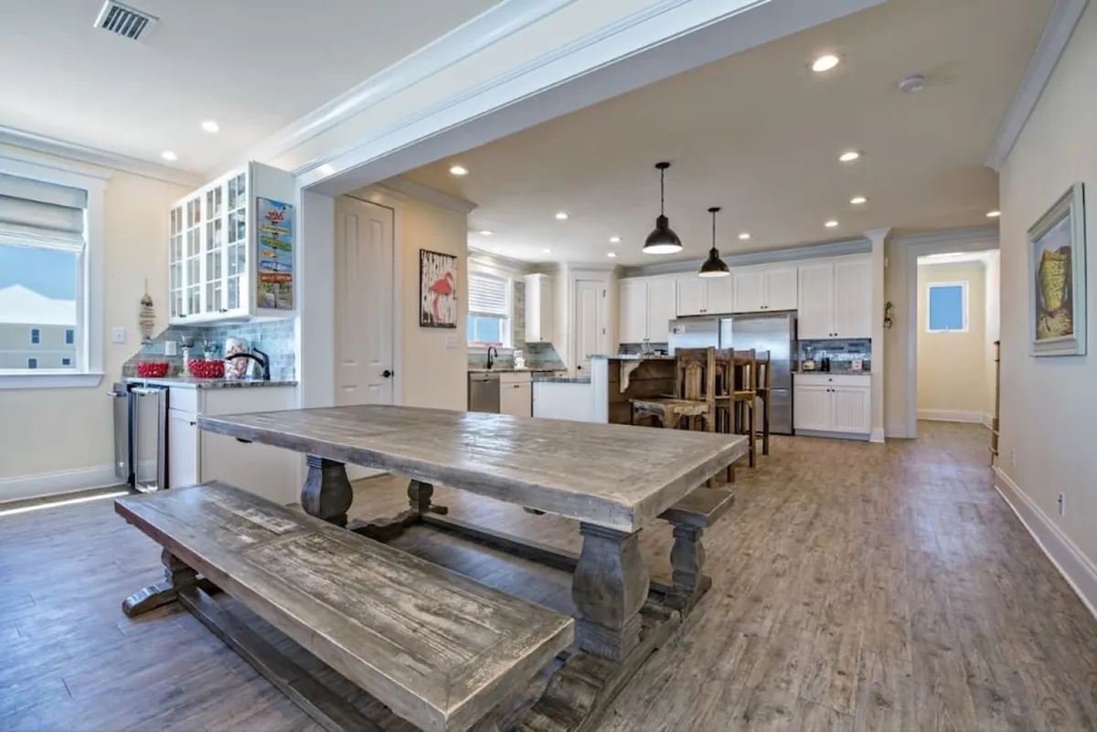 A warm, inviting open-plan dining and kitchen space with a large rustic wooden dining table and bench seating, modern white cabinetry with stainless steel appliances, and a kitchen island with wooden bar stools. Decorative pendant lights above the island add a touch of charm.