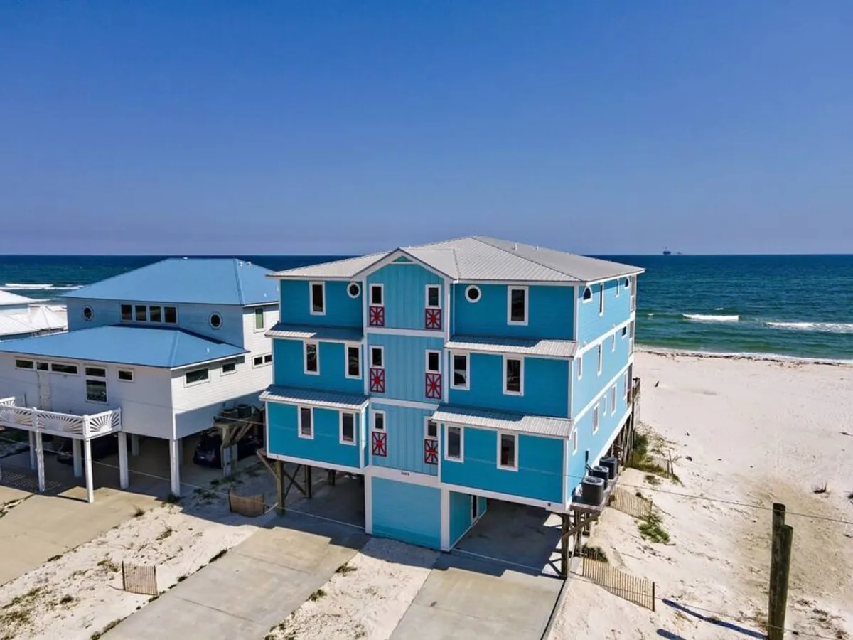 This image displays a bright blue duplex beach house on the sugar-white sand, set against the clear blue ocean and sky. The property’s spacious decks and proximity to the water are evident, and the duplex structure suggests the option to rent both sides for larger groups. The overall scene conveys tranquility and exclusivity.