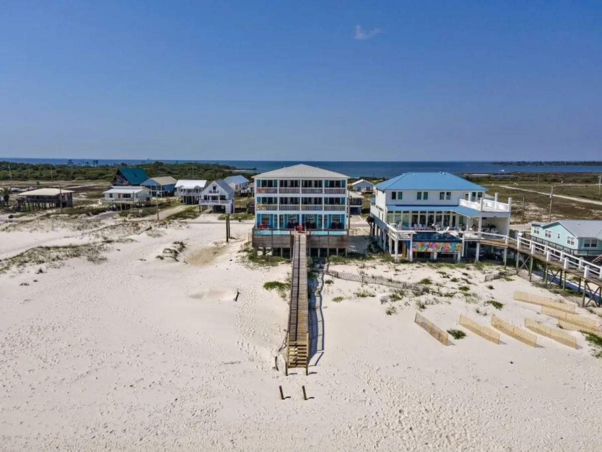 A large three-story beach house with expansive balcony and deck areas sits at the end of a long wooden walkway, extending over pristine white sand towards the blue Gulf waters. The scene includes adjacent beach houses with colorful roofs, and a wide, uncrowded stretch of beach.