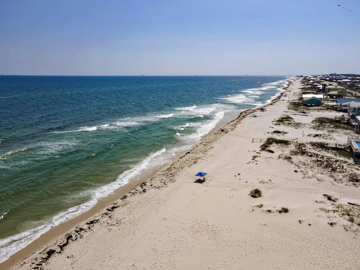 A quiet, endless stretch of white sandy beach meets the blue Gulf of Mexico waters. A single blue umbrella sits on the sand, while a line of beachfront houses can be seen in the distance. The beach is peaceful and inviting, perfect for relaxation or recreation.