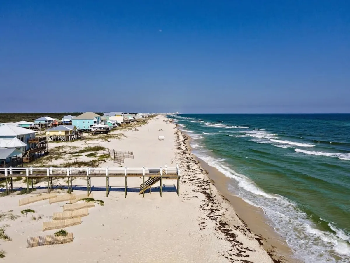 A long stretch of sugar-white sandy beach bordered by gently rolling ocean waves. Elevated beach houses line the shore, and a wooden walkway leads visitors directly from the houses to the secluded beach. The scene is peaceful and inviting, with an expansive view of both the sand and the Gulf horizon.