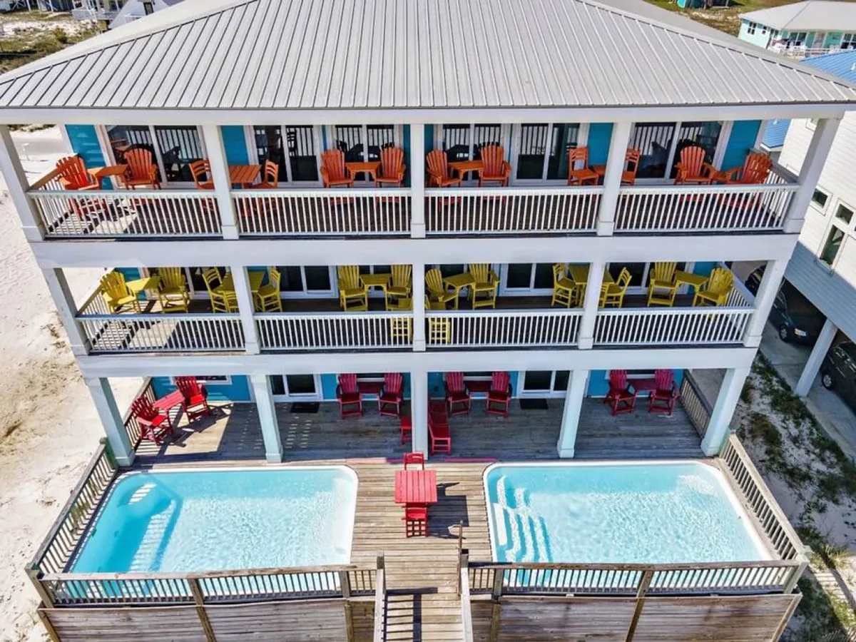 This image shows a vibrant exterior view of a beach house featuring two private swimming pools, a large wooden deck with ample seating provided by colorful Adirondack chairs, and a three-level balcony structure. The setting is cheerful and clearly designed for group enjoyment and gatherings, with multiple spaces for outdoor activities.
