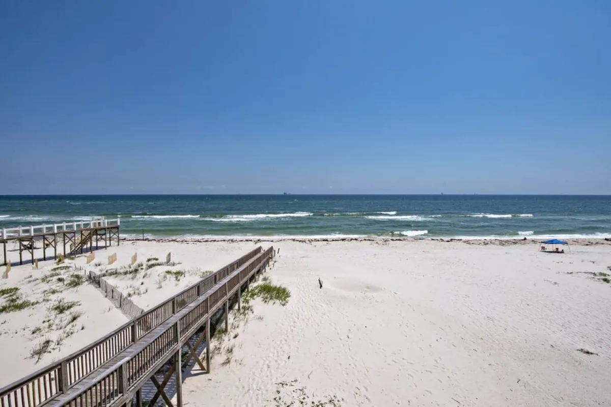 A wooden boardwalk stretches over pristine white sand dunes, leading directly to the calm, inviting Gulf of Mexico. Gentle waves roll onto the shore under a clear blue sky, and the untouched landscape exudes seclusion and tranquility.