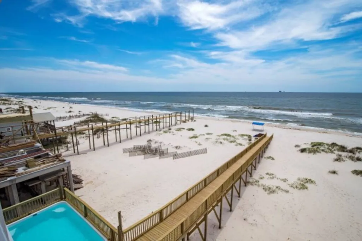 This image showcases a private beachfront pool with turquoise water, set next to a wooden boardwalk that leads directly to a pristine, sugar-white sandy beach. The calm Gulf of Mexico waves are visible just beyond the sand, and the pool area is surrounded by a sense of tranquility and seclusion.