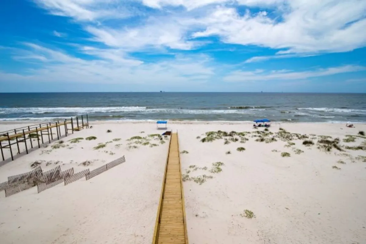 A weathered wooden boardwalk leads out to a quiet stretch of white sandy beach dotted with sparse vegetation. Two beach umbrellas offer shaded spots overlooking the blue ocean, with gentle waves and a wide, open sky creating a sense of escape.