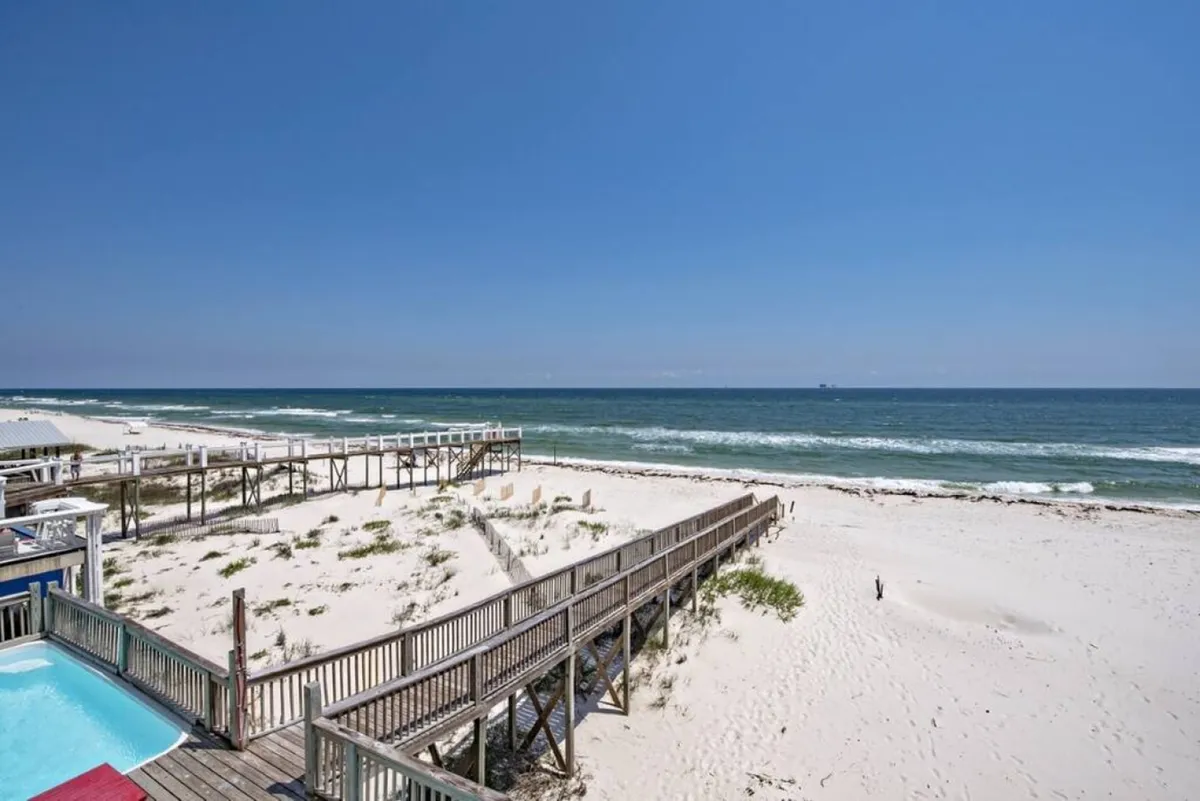 This exterior shot features a private pool right next to the beach house, with a wooden walkway leading directly to a white sandy beach and the calm Gulf waters beyond. The pool area is tranquil, with uninterrupted ocean views and a sense of seclusion.