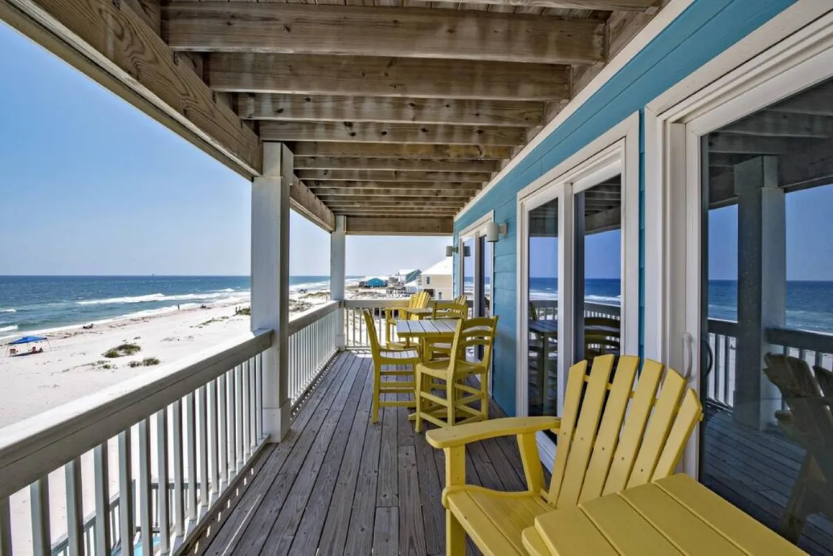 A wooden beachfront deck with bright yellow Adirondack chairs and a table set, overlooking expansive sugar-white sands and the Gulf of Mexico. The deck railing and overhead beams frame the stunning ocean view, inviting guests to relax and socialize outdoors.