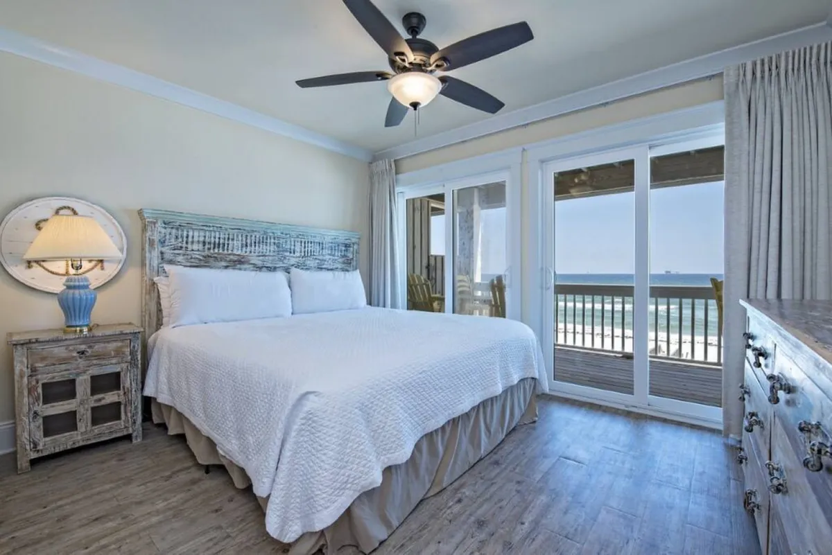 A king-sized bed dressed in crisp white linens is set against a backdrop of large sliding glass doors, which open onto a balcony overlooking the ocean. Rustic wooden furniture, including a nightstand and dresser, complement the neutral palette, while natural light fills the room, evoking a restful, spa-like ambiance.
