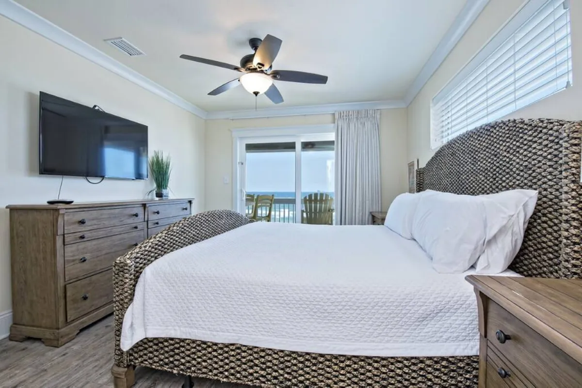 A spacious coastal bedroom features a large woven textured bed, a wooden dresser with a flat-screen TV, and a sliding glass door leading to a balcony overlooking the ocean. The aesthetic is restful and inviting, with gentle light and a ceiling fan for comfort.