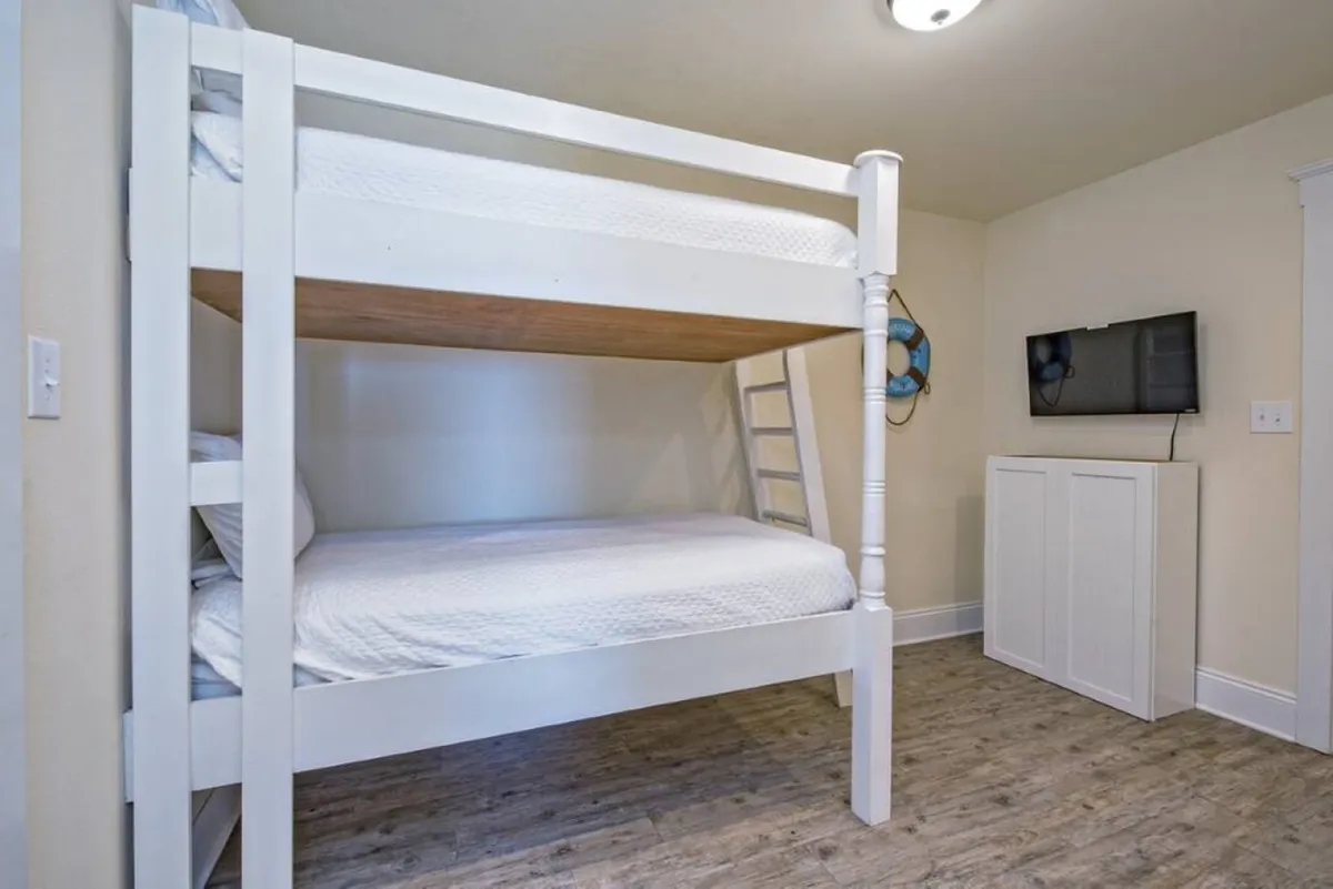 A bright, family-friendly bunk room features a white wooden bunk bed with a twin-over-full design, wall-mounted TV, nautical life preserver decor, and wood-style flooring. The space is welcoming and practical, ideal for kids or groups, with ample space for play and relaxation.