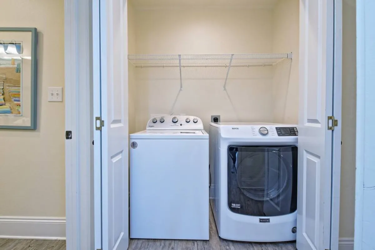 A dedicated laundry utility area with a white top-loading washing machine and front-loading dryer, both tucked neatly behind open bi-fold doors. A wire shelf provides extra storage, creating a practical, organized space for handling laundry and beach gear with ease.