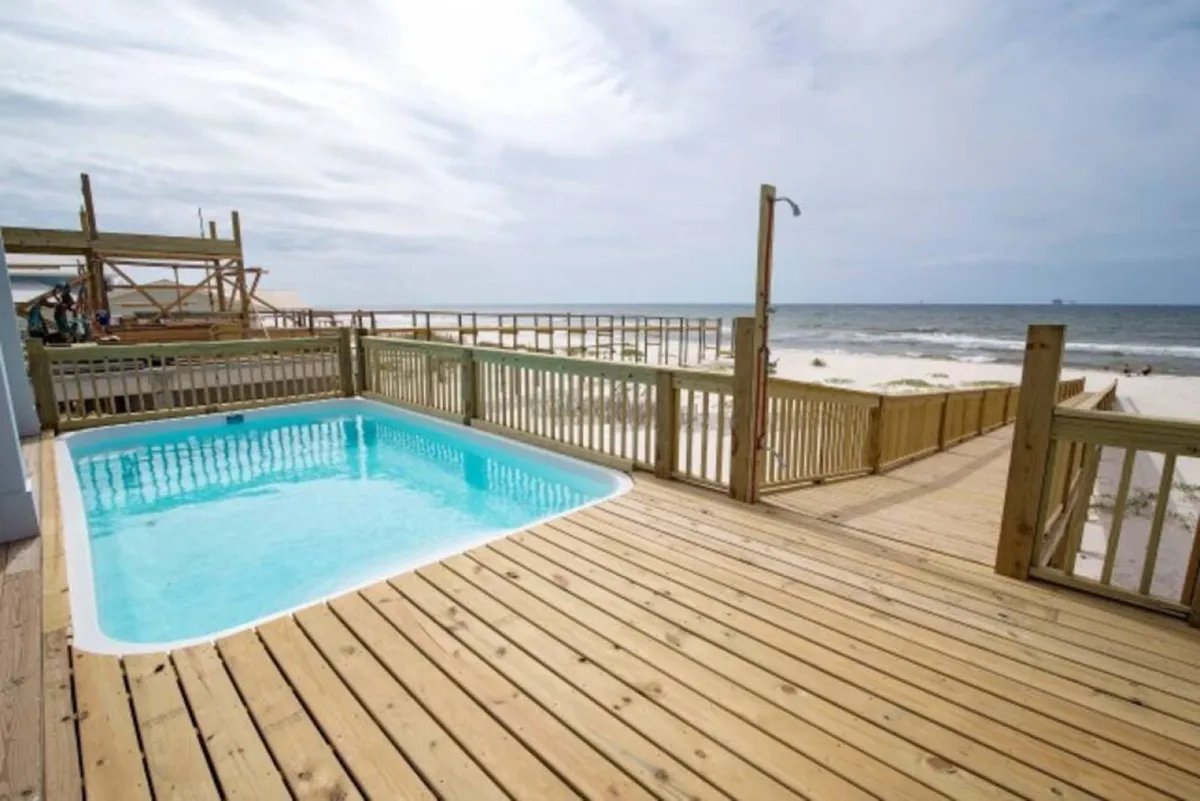 A sparkling private swimming pool with clear blue water sits just off a wooden deck, which leads toward the sandy beach and ocean beyond. The space offers both poolside lounging and immediate access to the Gulf, epitomizing luxurious beachfront living.