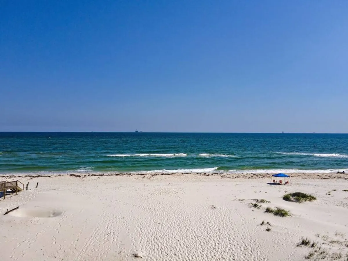 A tranquil, sugar-white sandy beach with calm Gulf waters, a small blue umbrella and chairs set up for relaxation, and natural dune vegetation scattered along the shore. The beach appears quiet and untouched, perfect for family picnics, shell hunting, or simply enjoying the sunrise.
